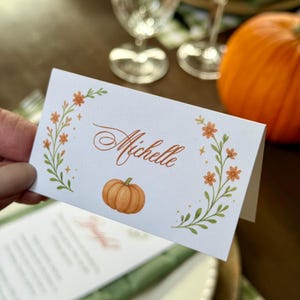 K&ouml;nnte beinhalten: Eine wei&szlig;e Tischkarte mit dem Namen "Michelle" in brauner Schrift, verziert mit einem Aquarellk&uuml;rbis und floralen Akzenten in Orange und Gr&uuml;n. Die gefaltete Karte liegt auf einem Tisch und deutet auf ein Herbst- oder Thanksgiving-Thema hin.