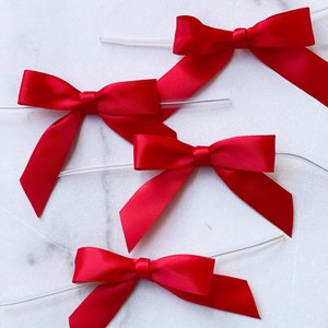 May include: Four red satin ribbon bows tied with clear string. The bows are arranged in a semi-circle on a white surface.