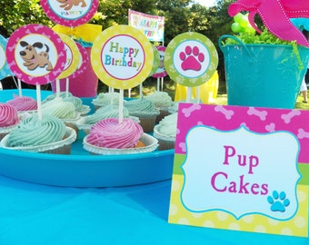 May include: A blue tray with colorful cupcakes decorated with pink and green frosting. A sign reads "Pup Cakes" with a paw print. The cupcakes are decorated with paw print toppers and "Happy Birthday" toppers.