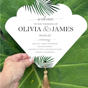 May include: A white wedding ceremony sign with a decorative palm leaf design and gold tassel. The sign reads "welcome" and announces the wedding of Olivia & James on June 28, 2023. Ceremony details are also listed.
