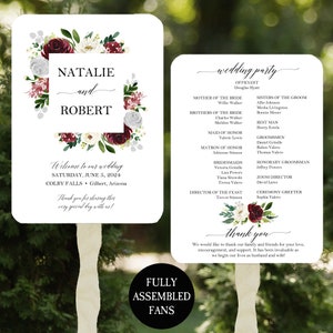 May include: Two white wedding program fans with wooden handles. The front features burgundy and white floral accents, the names Natalie and Robert, and event details. The back lists the wedding party members and a thank you message to guests.