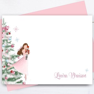 May include: A pink stationery set featuring a ballerina holding a nutcracker near a decorated Christmas tree. The set includes a white card with the name "Laura Braison" and a pink envelope. The illustration has a watercolor style with soft colors and snowflake accents.