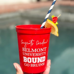May include: A red plastic cup with white text that reads "Congrats Caroline! BELMONT UNIVERSITY BOUND GO BRUINS!". The cup has a blue and white striped straw, a lemon slice, and a decorative red rose. The cup is held in front of a blurred background.