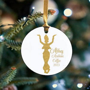 May include: White round Christmas ornament with a gold silhouette of a woman with her arms raised. The ornament has the text "Abbey Arabian Coffee 2023" in gold. A gold ribbon is attached for hanging.