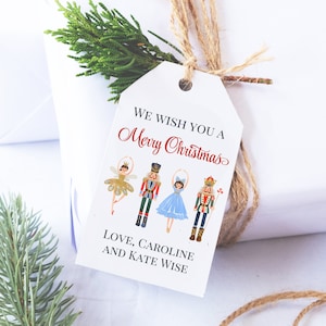 May include: A white gift tag with the text "We Wish You a Merry Christmas" in red script. The tag features illustrations of a ballerina and two nutcrackers. The tag is tied with twine and adorned with a sprig of greenery.