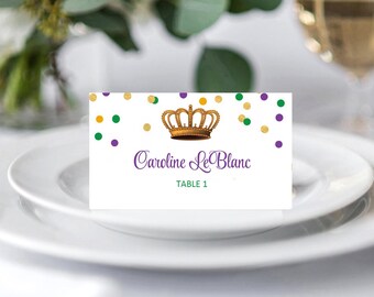 May include: A white place card with a gold crown illustration, purple text reading "Caroline LeBlanc" and "Table 1." The card is decorated with gold, green, and purple confetti. The card is on a white plate.