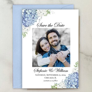 May include: A wedding invitation with a photo of a smiling couple, surrounded by blue and white floral accents. The text "Save the Date" is at the top, with the couple's names and wedding date below. The invitation is paired with a light blue envelope.
