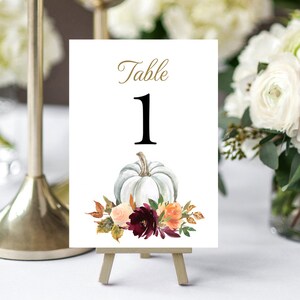 May include: A white table number card with the word "Table" in gold script above a large black number "1". A watercolor illustration of a white pumpkin and floral arrangement decorates the bottom of the card. The card is displayed on a small wooden easel.
