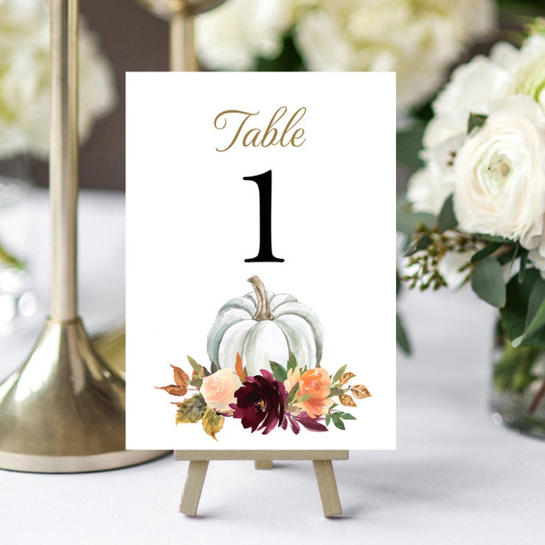May include: A white table number card with the word "Table" in gold script above a large black number "1". A watercolor illustration of a white pumpkin and floral arrangement decorates the bottom of the card. The card is displayed on a small wooden easel.