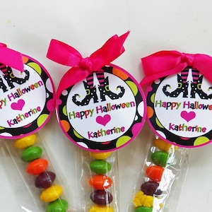 May include: Three Halloween-themed candy favors. Each favor features a clear cellophane bag filled with colorful candies, topped with a round tag. The tags read "Happy Halloween" and "Katherine" with witch legs and a pink bow.