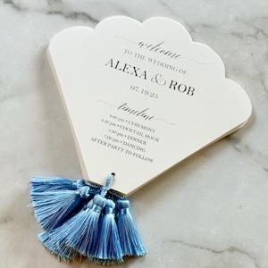 May include: A white wedding fan with a scalloped edge, featuring the text "welcome to the wedding of ALEXA & ROB" and a timeline for the event. The fan is adorned with a cluster of light blue tassels.
