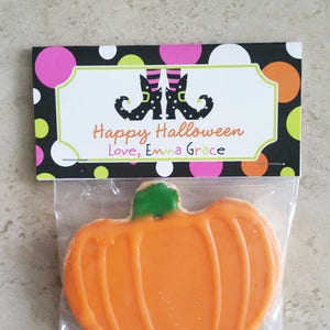 May include: A Halloween-themed treat bag with an orange pumpkin-shaped cookie. The bag has a decorative tag with the text "Happy Halloween" and witch boots. The tag also includes the text "Love, Emma Grace". The background is black with colorful polka dots.