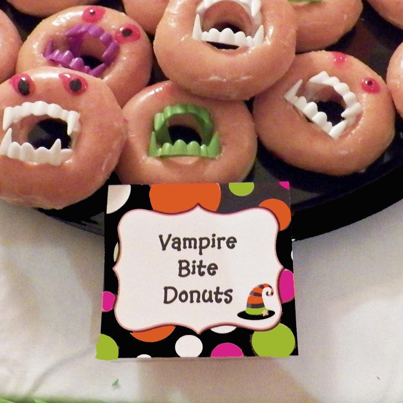 May include: A tray of Halloween-themed donuts with a vampire bite design. The donuts are glazed and decorated with plastic fangs and colorful accents. A sign reads "Vampire Bite Donuts" with a witch hat illustration.