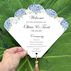 May include: A white wedding fan with a scalloped edge, adorned with blue and white floral accents. The fan features the text "Welcome" and details for Olivia & Trent's wedding ceremony, including the date and order of events. A gold tassel hangs from the bottom.