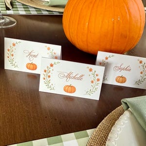 K&ouml;nnte beinhalten: Wei&szlig;e Tischkarten mit Namen in eleganter Schrift, verziert mit K&uuml;rbisillustrationen und floralen Akzenten. Im Hintergrund befindet sich ein gro&szlig;er orangefarbener K&uuml;rbis, der ein Herbst- oder Thanksgiving-Thema suggeriert. Die Karten liegen auf einem Holztisch.