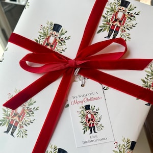 May include: White gift wrap with a red velvet ribbon and a gift tag that says "We wish you a Merry Christmas" with a nutcracker illustration. The gift wrap is decorated with nutcracker illustrations and green foliage.
