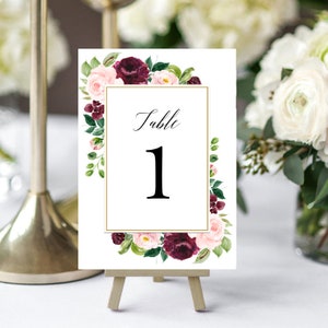 May include: White card with a gold frame and burgundy and pink floral border. The card says "Table 1" in black script.
