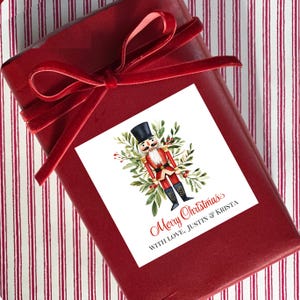 May include: A wrapped gift box with a red velvet ribbon. A white square sticker features a watercolor illustration of a nutcracker soldier surrounded by greenery. The sticker reads "Merry Christmas" and "With Love, Justin & Krista."