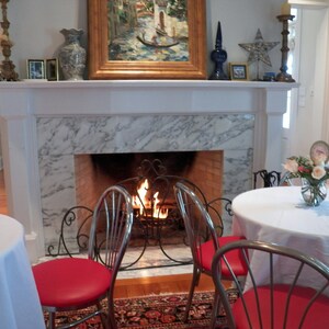 May include: A white marble fireplace with a fire burning inside. There are two white tables with white tablecloths and red chairs in front of the fireplace.