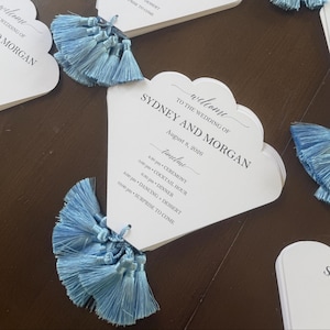 May include: A white fan-shaped wedding program card with scalloped edges, featuring black printed text that reads Welcome to the wedding of Sydney and Morgan, August 8, 2026, and a timeline of events, accented by a light blue silk tassel at the base.