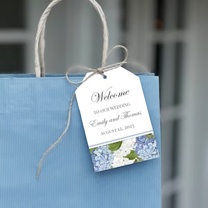 May include: A white wedding favor tag with blue hydrangeas and the text "Welcome to our wedding Emily and Thomas August 12, 2023".