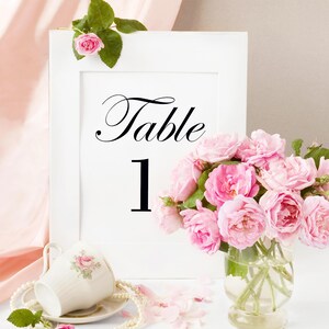 May include: White table number card with black text that reads "Table 1". Pink roses in a clear glass vase are in front of the card.