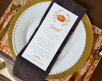 May include: Thanksgiving table setting with a white plate, gold charger, and brown napkin. A card with a pumpkin illustration and the word "Grateful" is on the napkin. Gold silverware and an orange patterned placemat complete the setting.