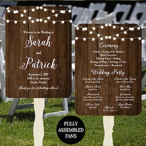 May include: Two wooden wedding fan signs with text. One says "Welcome to the Wedding of Sarah and Patrick" with the date and location. The other lists the ceremony details and wedding party. Both have string lights.
