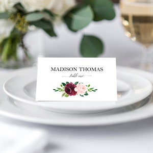May include: White place card with black text that reads "MADISON THOMAS - Table One" with a watercolor floral design of burgundy and pink roses with green leaves.