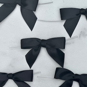 May include: Several black satin bows with clear plastic ties are arranged on a light gray surface. The bows are tied in a classic style, with neatly formed loops and tails. The satin has a slight sheen, and the bows are evenly spaced.