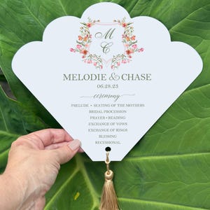 May include: A light blue wedding program fan with a floral crest and the names "Melodie & Chase" and the date "06.28.23". The program includes the ceremony details and has a gold tassel.