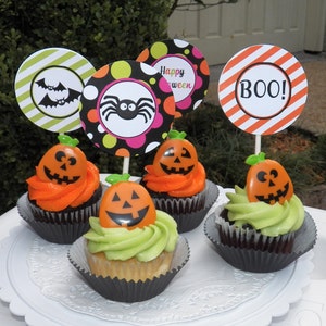 May include: A collection of Halloween cupcakes with orange and green frosting. Each cupcake is topped with a plastic jack-o'-lantern and a decorative paper circle with Halloween-themed designs and the word "BOO!"