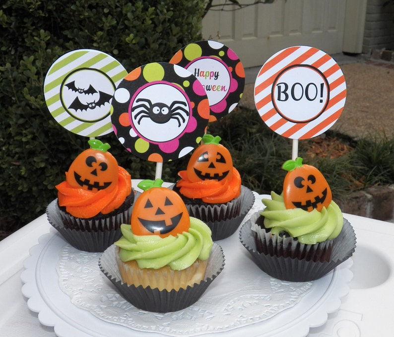 May include: A collection of Halloween cupcakes with orange and green frosting. Each cupcake is topped with a plastic jack-o'-lantern and a decorative paper circle with Halloween-themed designs and the word "BOO!"