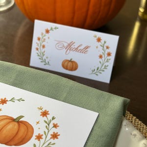 K&ouml;nnte beinhalten: Eine wei&szlig;e Tischkarte mit dem Namen "Michelle" in eleganter Schrift, umgeben von einem floralen Kranz mit orangefarbenen Bl&uuml;ten und einer K&uuml;rbisillustration. Eine gr&uuml;ne Serviette und ein gro&szlig;er orangefarbener K&uuml;rbis im Hintergrund.
