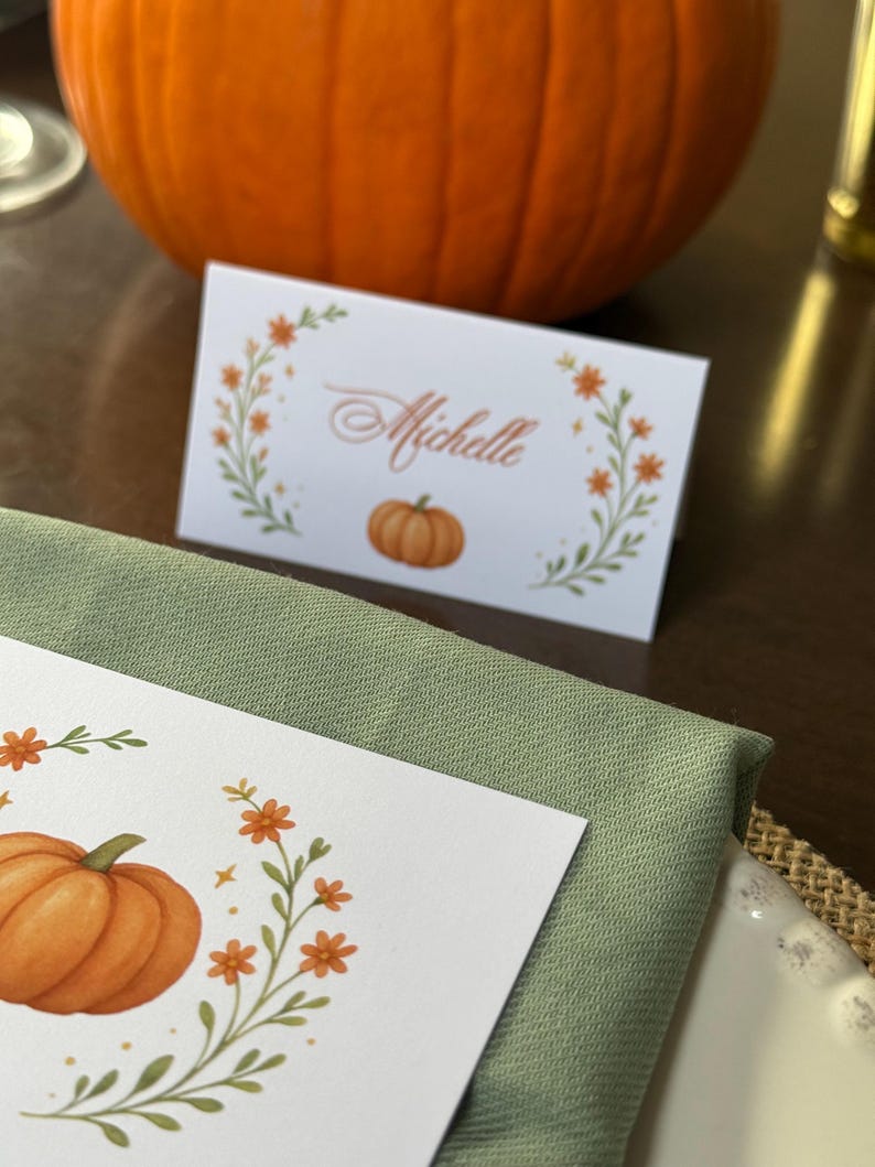 K&ouml;nnte beinhalten: Eine wei&szlig;e Tischkarte mit dem Namen "Michelle" in eleganter Schrift, umgeben von einem floralen Kranz mit orangefarbenen Bl&uuml;ten und einer K&uuml;rbisillustration. Eine gr&uuml;ne Serviette und ein gro&szlig;er orangefarbener K&uuml;rbis im Hintergrund.