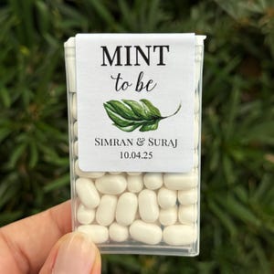 May include: A clear plastic box filled with white mints. The box has a white label that reads "MINT to be" with a green leaf illustration, names, and a date. The box is held in a person's hand.