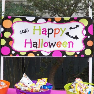 May include: A colorful Halloween banner with the words "Happy Halloween" in green, orange, and pink. The banner features a spider and bats, and is decorated with polka dots in various colors. Baskets of chips are visible below.