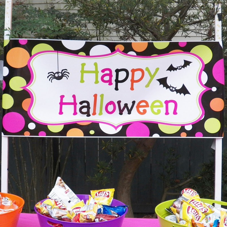 May include: A colorful Halloween banner with the words "Happy Halloween" in green, orange, and pink. The banner features a spider and bats, and is decorated with polka dots in various colors. Baskets of chips are visible below.