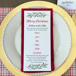 May include: A festive Christmas table setting with a gold charger, white plate, and red napkin. The napkin holds a card with the text "Merry Christmas" and a list of values. Gold cutlery and a plaid placemat complete the holiday theme.