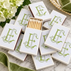 May include: A collection of rectangular matchboxes featuring a white cover with a decorative green letter E monogram. One open matchbox reveals a row of wooden matches with white tips. The boxes are arranged in a cluster on a surface.
