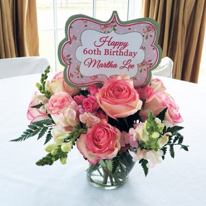 May include: A pink and green floral centerpiece with a sign that reads "Happy 60th Birthday Martha Lee".