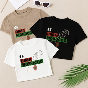 Puede incluir: Tres camisetas de manga corta en beige, negro y blanco. Cada camiseta muestra el texto "DIMA MOROCOO" en rojo y verde, un contorno de mapa y un emblema circular. Gafas de sol descansan encima.