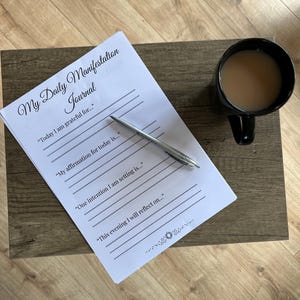 May include: A black mug filled with a light brown liquid sits on a wooden surface next to a white journal and a silver pen. The journal is titled "My Daily Manifestation Journal" and has prompts for gratitude, affirmations, and intentions.