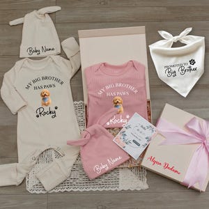 May include: A gift set for a new baby, including a cream-colored onesie and hat with the text "My Big Brother Has Paws" and "Rocky". A pink onesie and hat are also included, along with a bandana that says "Promoted to Big Brother". A gift box with a pink ribbon is also included.