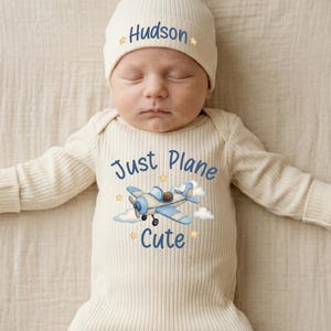 May include: A cream-colored ribbed long-sleeve onesie and matching beanie set. The beanie features the name Hudson in blue text with small stars. The onesie displays a blue airplane graphic, clouds, stars, and the text Just Plane Cute.