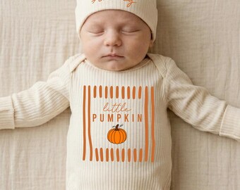 May include: A cream-colored ribbed long-sleeve onesie and matching beanie set. The beanie features the name Aubrey in orange script. The onesie displays the text little PUMPKIN in orange with a graphic of an orange pumpkin and vertical lines.