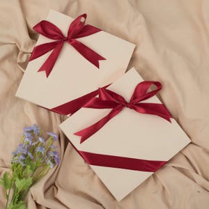 May include: Two cream-colored gift boxes with burgundy satin ribbons and bows. The boxes are arranged on a beige fabric surface, with a small bouquet of purple flowers in the lower left corner.