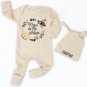 May include: A cream-colored baby onesie and matching hat set. The onesie features a bee-themed design with the text "New to the Hive". The hat has the word "name" printed on it. The set is made of a ribbed fabric.