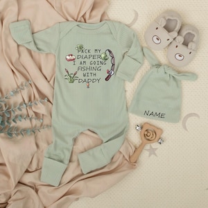 May include: A light green baby outfit set with a long-sleeved onesie, a knotted hat, and bear-shaped slippers. The onesie has the text "Pack my diaper I am going fishing with Daddy" and a fishing-themed graphic. A wooden rattle is also included.