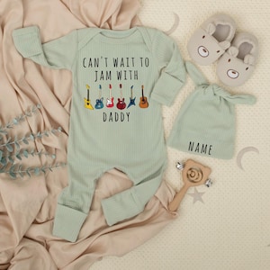 May include: A light green baby onesie with the text "CAN'T WAIT TO JAM WITH DADDY" and guitar graphics. The image also includes a matching hat with "NAME" printed on it, bear-shaped slippers, and a wooden rattle.